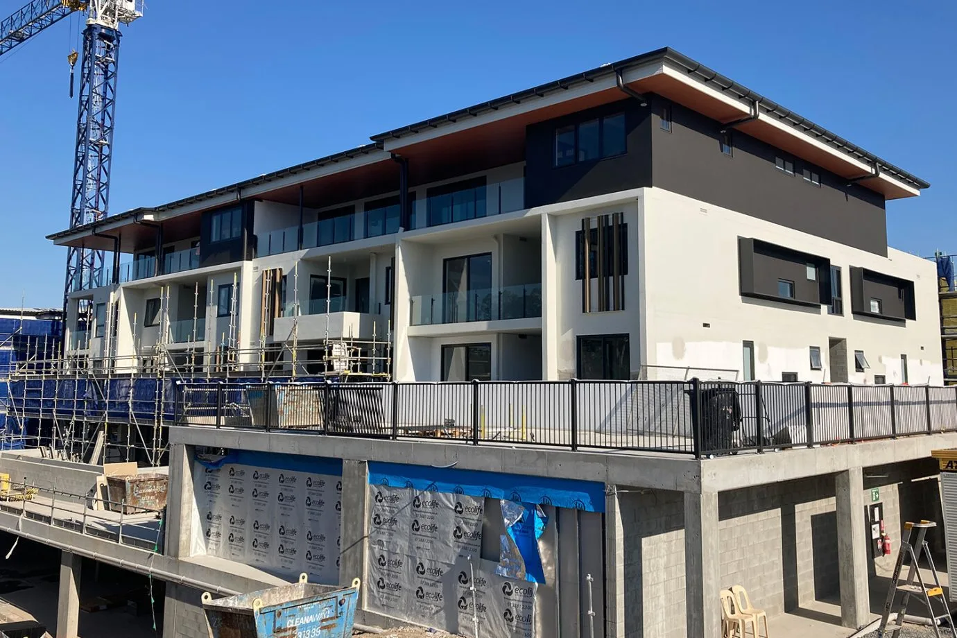 New apartment building with scaffolding and cladding on the second level. A crane can be seen from the corner and blue sky above.